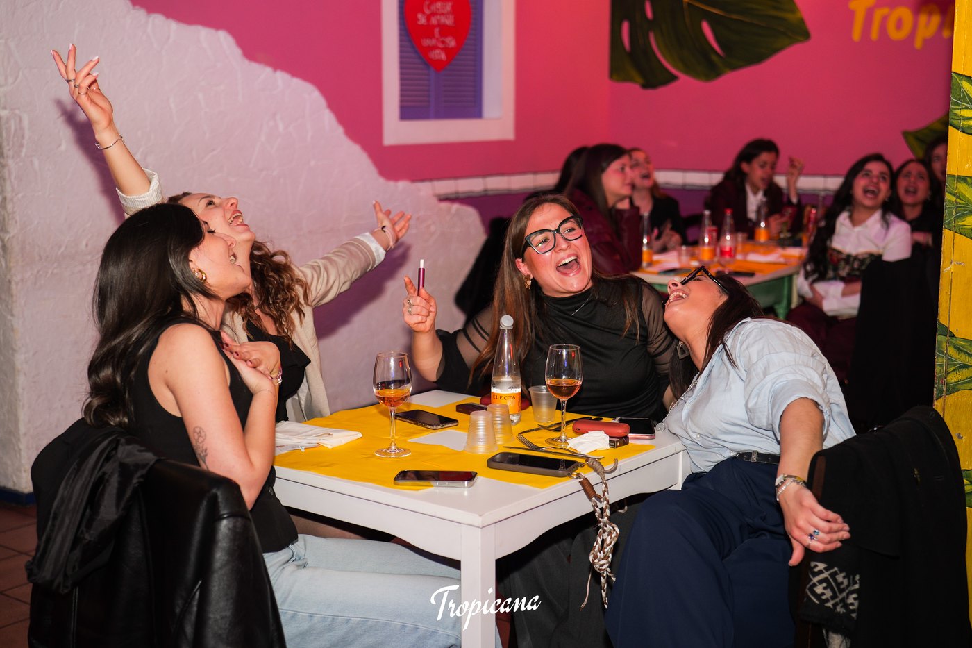 Amiche ridono e cantano a braccia alzate a un tavolo giallo del Tropicana, tra calici di vino e pareti rosa