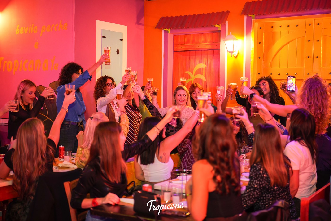 Donne brindano con boccali di birra a una lunga tavolata, pareti rosa e arancioni con scritta «bevila perché è Tropicana